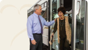 An older woman steps off a bus smiling, greeted by a man in a blue shirt and tie. She holds an umbrella and wears a fringe vest and necklace. The background shows the open door of a bus.