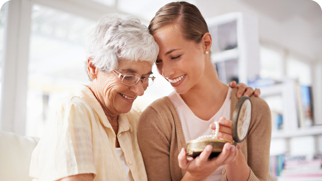 Elderly woman with short white hair and glasses smiles and hugs a young woman with a ponytail. Both are looking at an open round jewelry box. They are seated on a couch with a bright, blurred background.