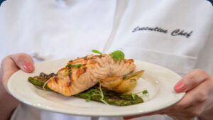 A person in a chef's coat holding a white plate with a grilled salmon fillet, roasted potatoes, asparagus, and microgreens garnish. The jacket has "Executive Chef" embroidered on it.