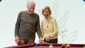 An elderly man and woman are playing pool, both smiling and enjoying the moment. The man is holding a cue stick, and the woman appears to be laughing joyfully. A soft, leafy pattern is in the background.