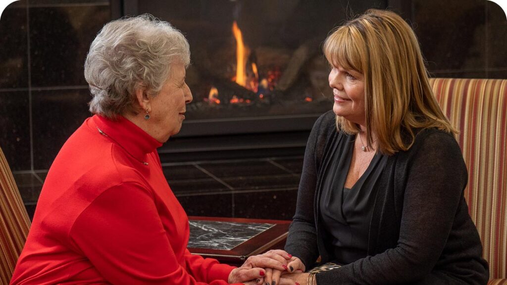 Two women sitting and holding hands in front of a fireplace. The woman on the left has short gray hair and wears a red top. The woman on the right has shoulder-length brown hair and wears a black outfit. They appear to be engaged in conversation.