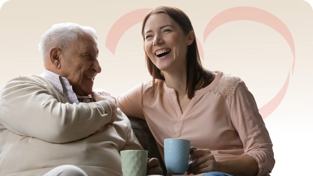 An elderly man and a young woman sit close together on a sofa, smiling and laughing while holding mugs. A soft heart shape is faintly visible in the background, symbolizing warmth and affection.