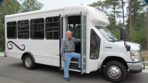 A man in a plaid shirt and blue jeans stands with one foot on the step of a parked white shuttle bus, holding the door open. Trees are visible in the background.