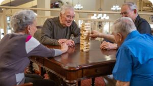 Four elderly individuals are sitting around a wooden table, playing a game of Jenga. They are engaged and smiling, with one person carefully pulling a block. The setting appears cozy, with warm lighting and soft furnishings.