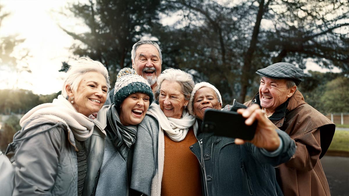 A group of six smiling older adults, dressed in winter clothing, stand close together outdoors as one takes a selfie. The background shows trees and a cloudy sky.