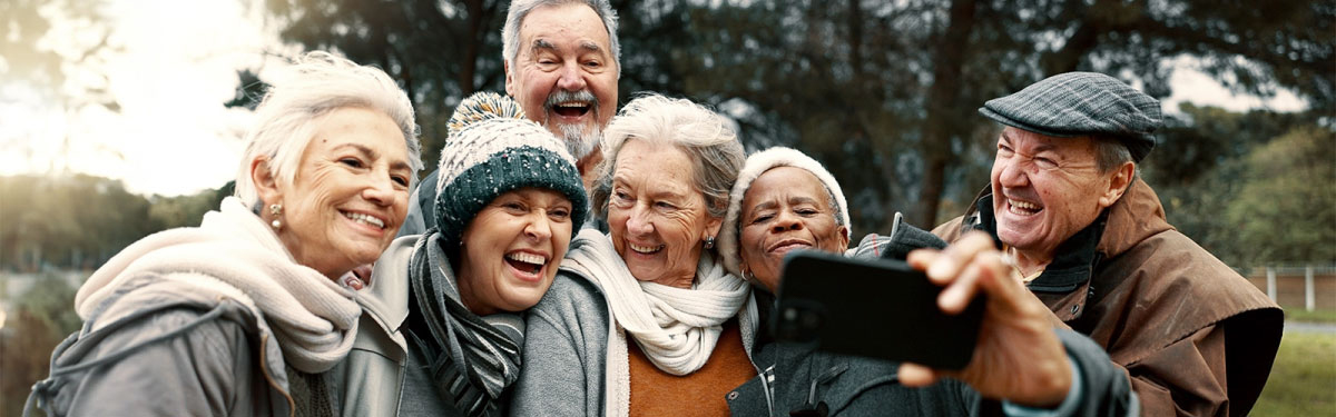 A group of six older adults bundled in winter clothes stand close together outdoors, smiling and laughing as one person holds a smartphone to take a selfie. Trees and soft sunlight are visible in the background.