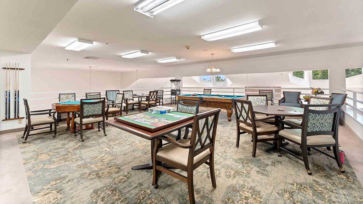 A bright recreation room with card tables, cushioned chairs, billiard tables, and pool cues. The space has white walls, overhead fluorescent lights, and a patterned carpet. Large windows provide natural light.