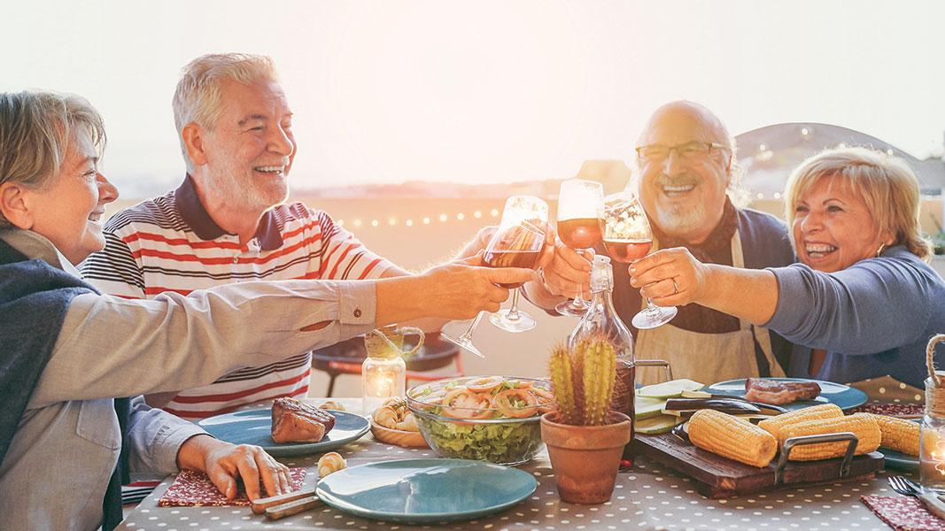 Four smiling older adults sit at an outdoor table, toasting with glasses of wine. The table is set with various foods including corn, salad, and steak, creating a cheerful and festive atmosphere at sunset.