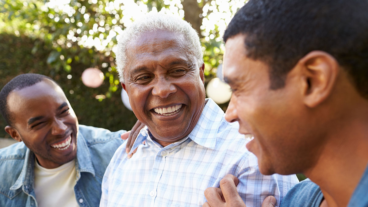 Three men, two younger and one older with gray hair, smile and laugh together outdoors on a sunny day, appearing happy and relaxed. The background is filled with greenery and soft sunlight.