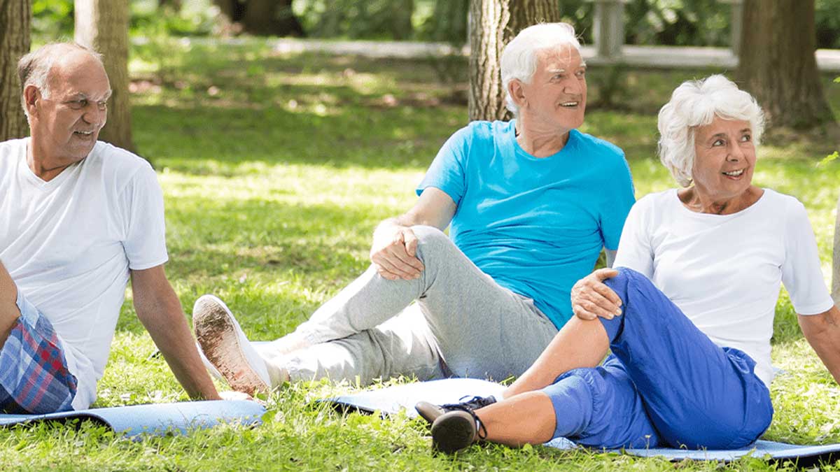 Three older adults sit on yoga mats outdoors, stretching and smiling while enjoying a sunny day in a park with green grass and trees in the background.