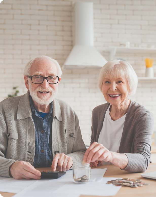 An elderly man and woman sit at a table with coins, a jar, and papers, smiling at the camera. The man uses a calculator while the woman holds the jar, in a bright, modern kitchen setting.