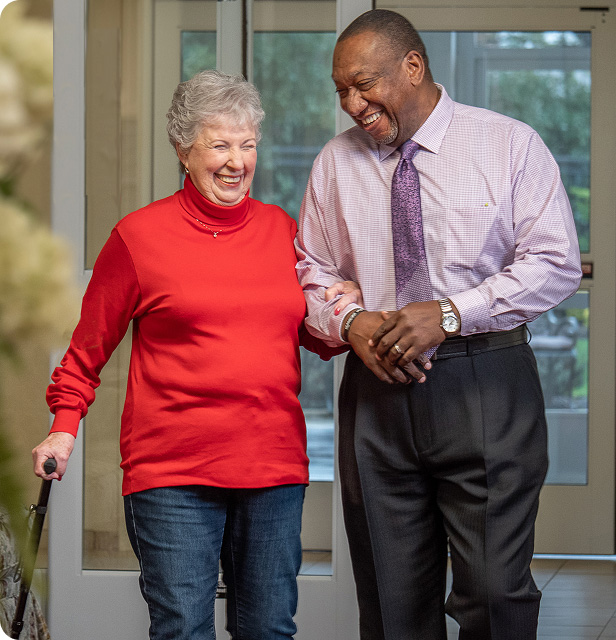 An older woman in a red sweater uses a cane and walks arm-in-arm with a man in a dress shirt and tie. Both are smiling and laughing as they walk indoors.