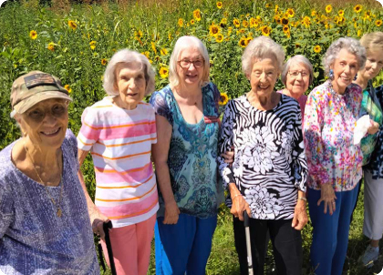 A group of elderly women stand outdoors in front of blooming sunflowers, smiling and posing for the photo on a sunny day. Some use canes for support.