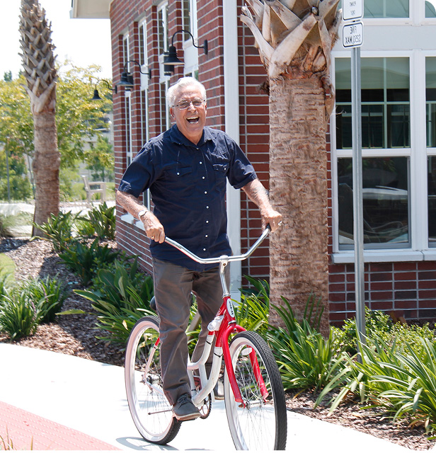 An older man with gray hair and glasses smiles as he rides a red and white bicycle on a sidewalk next to a red brick building, with palm trees and greenery in the background.
