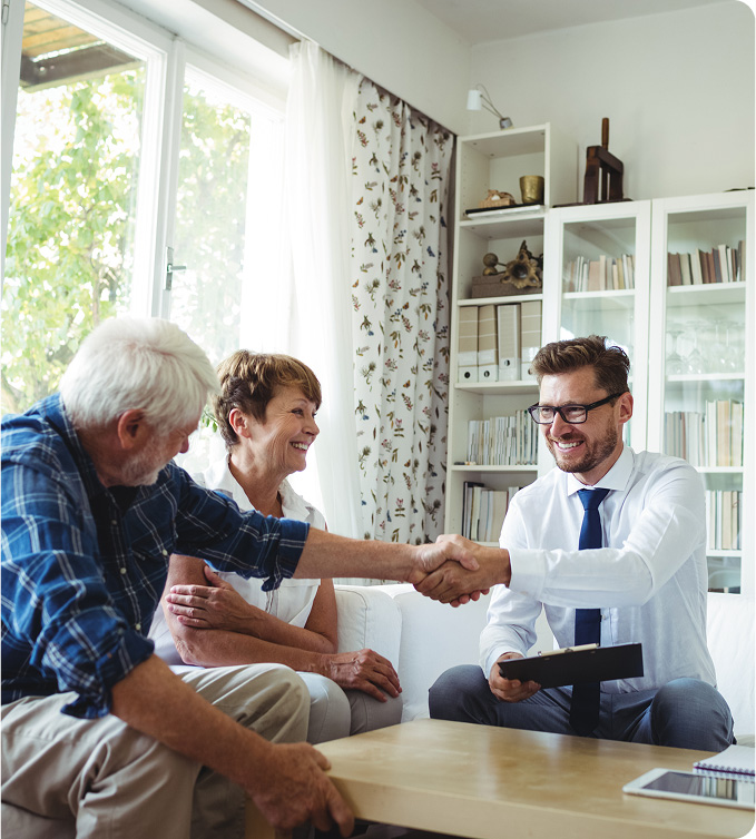 A smiling older couple sits on a couch in a bright living room, shaking hands with a young man in glasses and a tie who is holding a clipboard, suggesting a friendly business meeting or consultation.