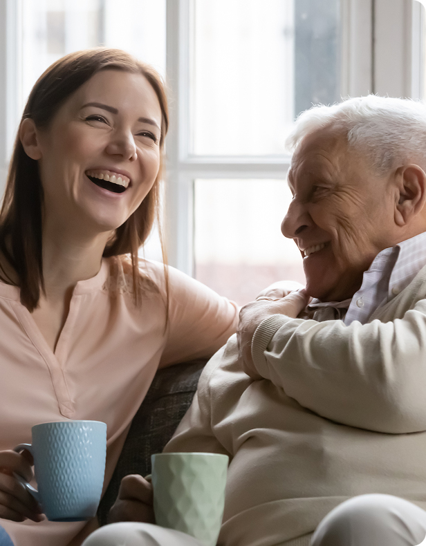 A young woman and an older man sit together on a couch, smiling and laughing while holding coffee mugs, with a bright window in the background.