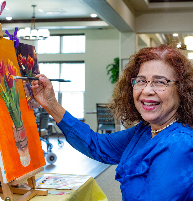 An older woman wearing glasses and a bright blue blouse smiles while painting colorful flowers on a canvas at an easel in a well-lit room.
