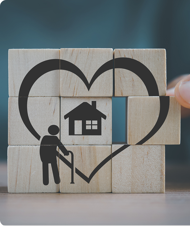 A hand arranges wooden blocks with icons of a heart, a house, and an elderly person using a cane, symbolizing elderly care or assisted living at home.