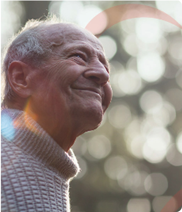 An elderly person with short gray hair smiles gently while looking up. They wear a light knit sweater, and soft sunlight with bokeh effects fills the blurry background.