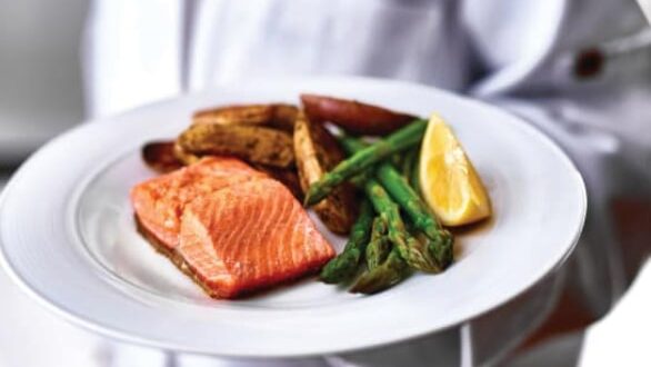A person in a white chef's uniform holds a plate with a serving of grilled salmon, roasted fingerling potatoes, asparagus, and a lemon wedge. The food is presented in an appetizing and detailed manner.