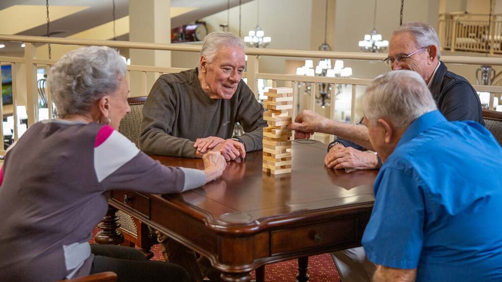 Four older adults sit around a wooden table, smiling and playing a game of Jenga together in a well-lit, spacious indoor setting.